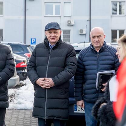 Trzy mężczyźni w zimowym stroju stoją przed budynkiem w parkingu, uczestniczą w transferze ambulansów do szpitali w Częstochowie i Kłobucku. (wygenerowano przez AI)