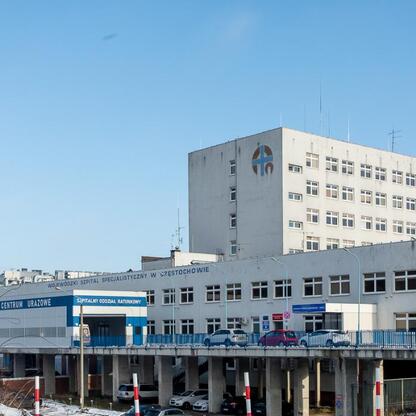 Wojewódzki Szpital Specjalistyczny w Częstochowie, ambulanse z Katowic, Trauma Center, Szpitalny Oddział Ratunkowy, niebieski dach, żółte logo, szare fasady, błękitne niebo, parkujące samochody. (wygenerowano przez AI)