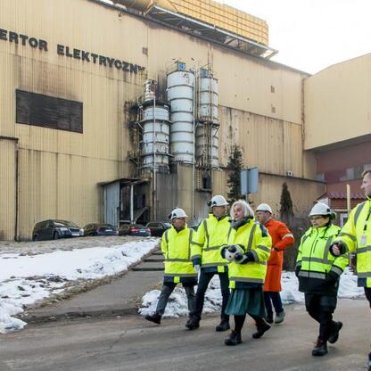 Grupa osób w kurtkach odblaskowych i hełmach spaceruje po terenie Huty Częstochowa, podczas inspekcji lub touru. (wygenerowano przez AI)