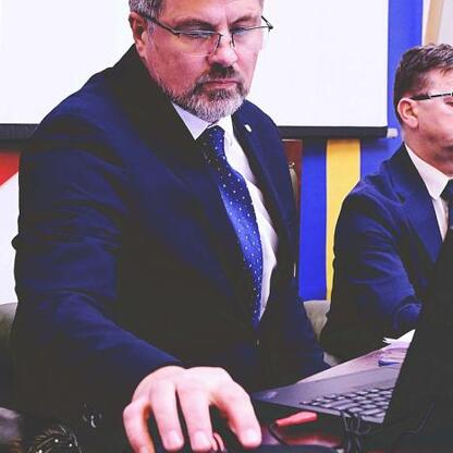 Mężczyzna w garniturze siedzi przy biurku, korzystając z komputera, w tle druga osoba w podobnym stroju. (wygenerowano przez AI)