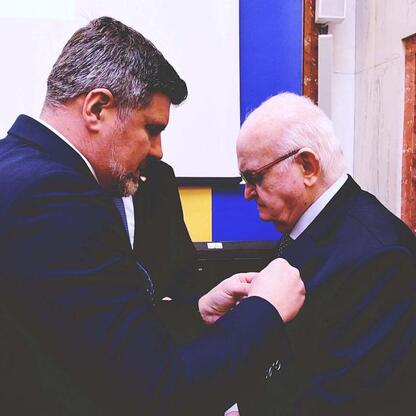 Dwóch mężczyzn w strojach formalnych, jednego z nich nagradzają lub prezentują coś drugiemu, prawdopodobnie medal lub certyfikat, w formalnym kontekście Sejmiku Województwa Śląskiego. (wygenerowano przez AI)