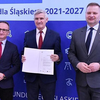 Trzech mężczyzn w strojach formalnych stojących przed niebieskim tłem z napisem 