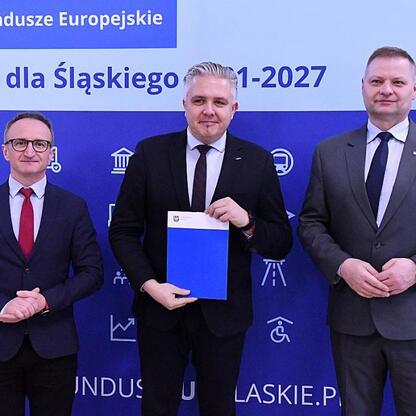 Trzech mężczyzn w strojach formalnych prezentuje dokumenty przed tłem z napisem 