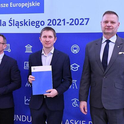 Trzech mężczyzn w strojach formalnych stojących przed niebieskim tłem z napisem 