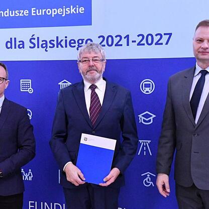 Trzech mężczyzn w strojach formalnych stoi przed tłem z napisem 