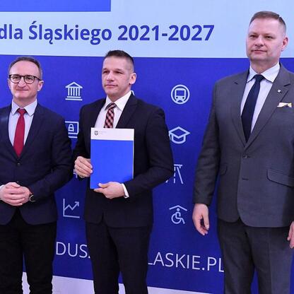 Trzech członków zarządu w formalnym stroju prezentuje dokumenty przed niebieskim tłem z ikonami i napisami związany z programem Funduszy Europejskich dla Śląska. (wygenerowano przez AI)