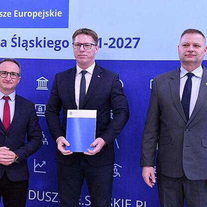 Trzech urzędników w strojach formalnych stojących przed tłem z napisem 