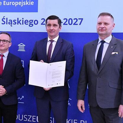 Trzech mężczyzn w strojach formalnych stoi przed tłem z napisem 