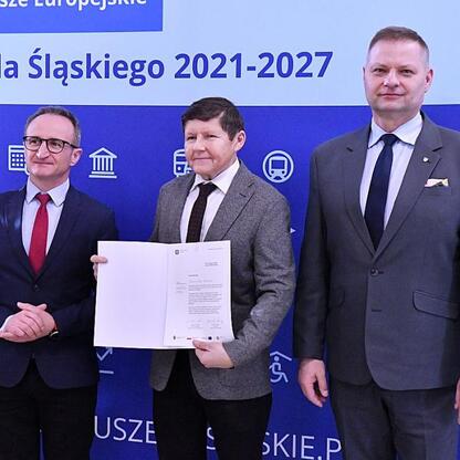 Trzech mężczyzn w strojach formalnych odbierają certyfikaty przed niebieskim tłem z ikonami i tekstem o funduszach dla projektów odnawialnych źródeł energii w Śląsku. (wygenerowano przez AI)