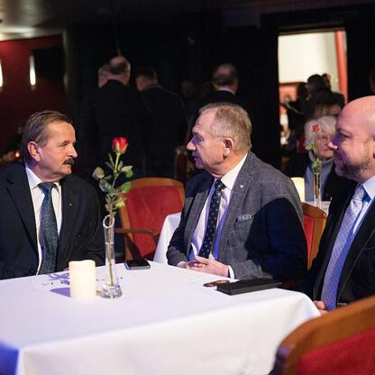 Formalne spotkanie w sali konferencyjnej, gdzie trzech mężczyzn w strojach formalnych rozmawiają przy stole z czerwonymi krzesłami i białymi obrusami. (wygenerowano przez AI)