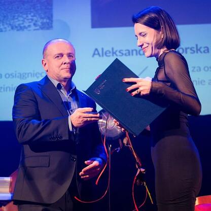 Podczas uroczystości wręczenia nagród Marszałka Województwa Śląskiego, mężczyzna w ciemnym garniturze prezentuje nagrodę kobiecie w czarnej sukni. (wygenerowano przez AI)