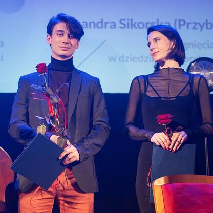 Na zdjęciu młody mężczyzna w czarnej kamizelce i pomarańczowych spodniach trzyma certyfikat i czerwony kwiat, stojąc na scenie podczas uroczystości wręczenia nagród kulturalnych dla Województwa Śląski (wygenerowano przez AI)