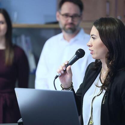 Podczas konferencji w Katowickim Centrum Onkologii, kobieta w czarnym płaszczu i białej bluzie przemawia do mikrofonu, podczas gdy w tle siedzą dwie osoby. (wygenerowano przez AI)