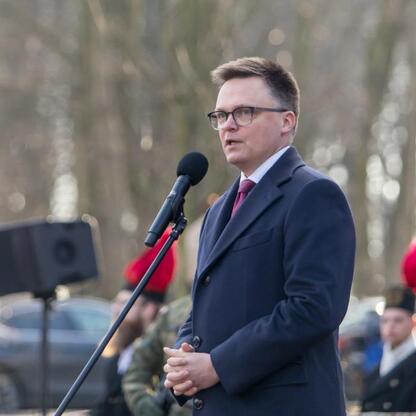 Mężczyzna w ciemnym garniturze i krawacie przemawia do mikrofonu na podium, podczas uroczystości upamiętniających 80. rocznicę Tragedii Górnośląskiej. (wygenerowano przez AI)