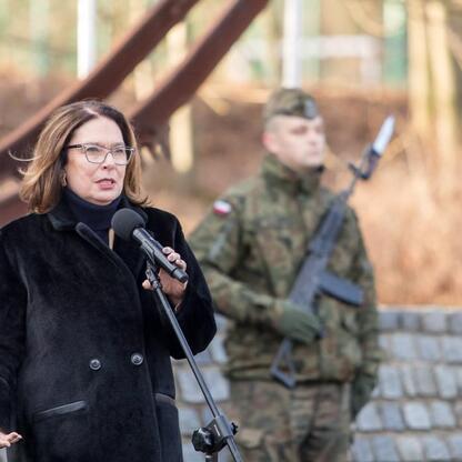 W czasie uroczystości upamiętniających 80. rocznicę Tragedii Górnośląskiej, kobieta w czarnej kurtce przemawia z podium, podczas gdy żołnierz w kamuflażu stoi za nią, trzymając karabin, symbolizując o (wygenerowano przez AI)