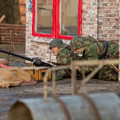 Rekonstrukcja wydarzeń z 1945 r. przedstawia żołnierzy w pozycji obronnej, z bronią i sprzętem wojskowym, przed ceglanym budynkiem z czerwonymi oknami. (wygenerowano przez AI)