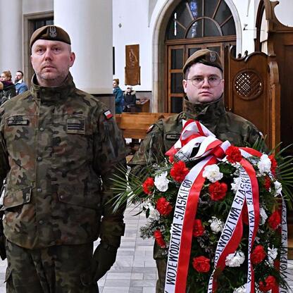 Dwóch żołnierzy w mundurach stojących w kościele, trzymających wieniec z czerwonymi i białymi wstążkami i białymi kwiatami, uczestniczących w uroczystościach upamiętniających 80. rocznicę Tragedii Gór (wygenerowano przez AI)