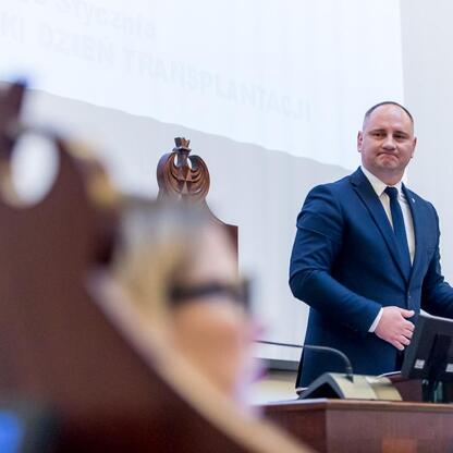 Na zdjęciu widoczny jest mąż w ciemnym garniturze, który przemawia z podium, prawdopodobnie na sesji Sejmiku Osób z Niepełnosprawnością Województwa Śląskiego. (wygenerowano przez AI)