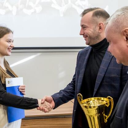 Na zdjęciu młoda kobieta odbiera nagrodę z rąk mężczyzny w garniturze, podczas uroczystości sportowej w Silesia. (wygenerowano przez AI)