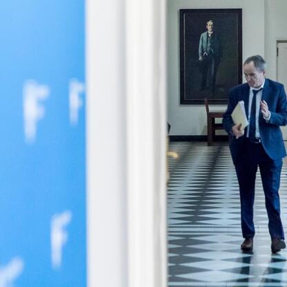 Mężczyzna w garniturze spaceruje po korytarzu, trzymając folder i gestykulując. W tle wiszący portret w garniturze. (wygenerowano przez AI)