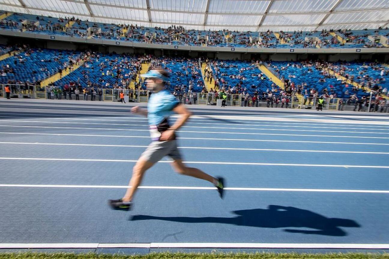 Zdjęcie przedstawia dynamiczną scenerię na stadionie, gdzie biegacz jest w pełnym biegu na niebieskim torze, z widocznymi kibicami w tle. (wygenerowano przez AI)