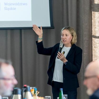 Podczas IV posiedzenia Rady ds. Sprawiedliwej Transformacji Województwa Śląskiego w Dąbrowie Górniczej kobieta prowadzi prezentację, gestykulując i trzymając mikrofon. (wygenerowano przez AI)