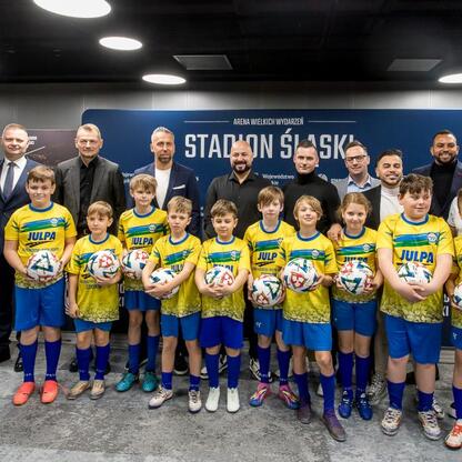 Grupa młodych piłkarzy w żółto-niebieskich strojach, trzymających piłki, pozują do zdjęcia przed Stadionem Śląskim. (wygenerowano przez AI)
