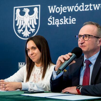 Wicemarszałek Województwa Śląskiego Grzegorz Boski przemawia na konferencji prasowej w Silesia, wspierany przez dwie damy. (wygenerowano przez AI)