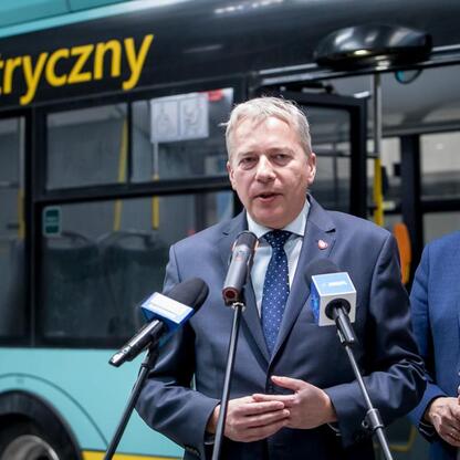 Marszałek Województwa Śląskiego Wojciech Saługa przemawia na tle elektrycznego autobusu, podkreślając ekologiczne i ekonomiczne zalety nowoczesnych pojazdów. (wygenerowano przez AI)