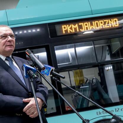 Mężczyzna przemawia na podium przed nowym elektrycznym autobusem PKM Jaworzno, przedstawiając program modernizacji komunikacji miejskiej. (wygenerowano przez AI)