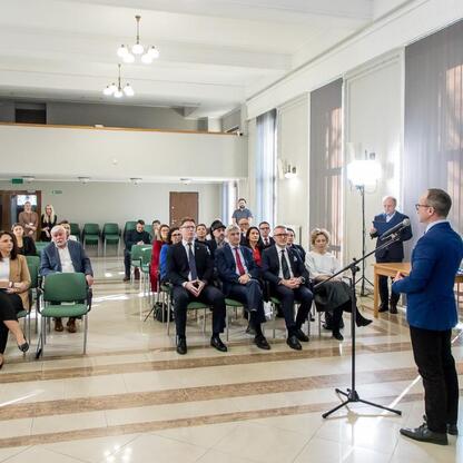 Formalne spotkanie w Silesia, ludzie w strojach formalnych siedzą w rzędach, słuchając mówcy stojącego na podium. (wygenerowano przez AI)