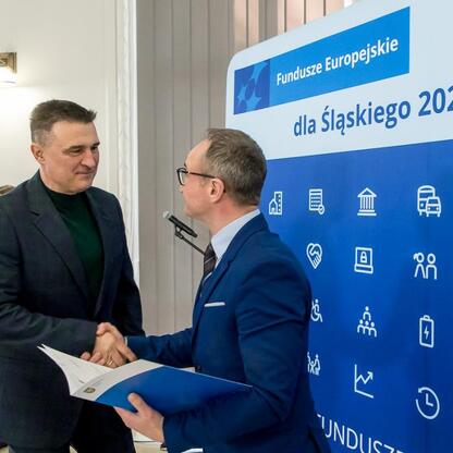 Dwóch mężczyzn w strojach formalnych trzyma się za ręce przed logo i napisami Funduszy Europejskich dla Śląska 2021-2027. (wygenerowano przez AI)