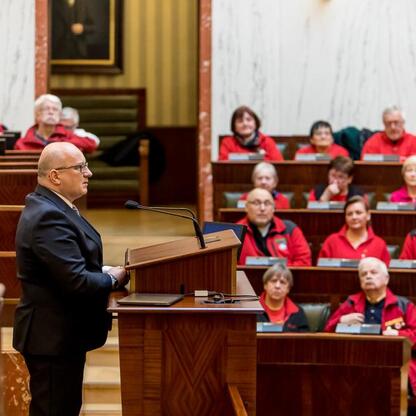 Formalna uroczystość w Sali Sejmu Śląskiego, gdzie przemawiający w garniturze adresuje grupę ludzi w czerwonych kamizelkach. Atmosfera formalna i uczciwa. (wygenerowano przez AI)