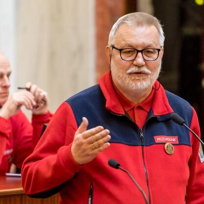 W Sali Sejmu Śląskiego uczczono 150-lecie przewodnictwa turystycznego. Przewodnicy w czerwono-niebieskich kurtkach przemawiają i notują, uczczeni przez Marszałka Województwa Śląskiego. (wygenerowano przez AI)