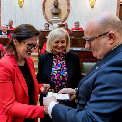 Formalna uroczystość w Sali Sejmu Śląskiego, gdzie kobieta w czerwonej kurtce otrzymuje nagrodę od mężczyzny w ciemnym garniturze. (wygenerowano przez AI)