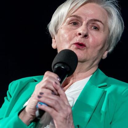 Kobieta w zielonym płaszczu przemawia w mikrofonie, uczestnicząc w dyskusji na konferencji o równości płci i współpracy. (wygenerowano przez AI)