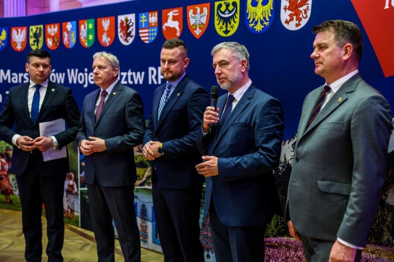 Mężczyzna w garniturze podpisuje dokument, podczas gdy druga osoba stoi za nim. Tło zaznacza Konwent Marszałków Małopolska 2023. (wygenerowano przez AI)