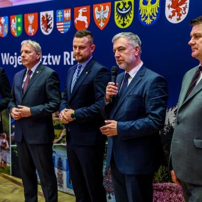 Grupa pięciu mężczyzn w strojach formalnych stoi przed tłem z herbami województw polskich i napisem 