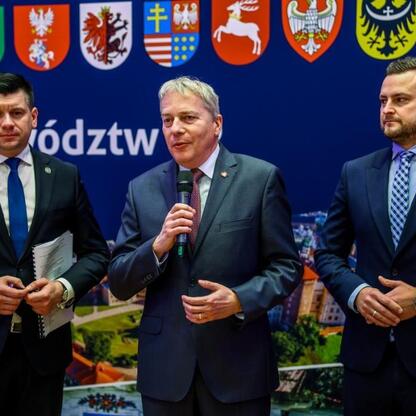Trzech mężczyzn w garniturach przedstawiających regionalne emblematy i napis 