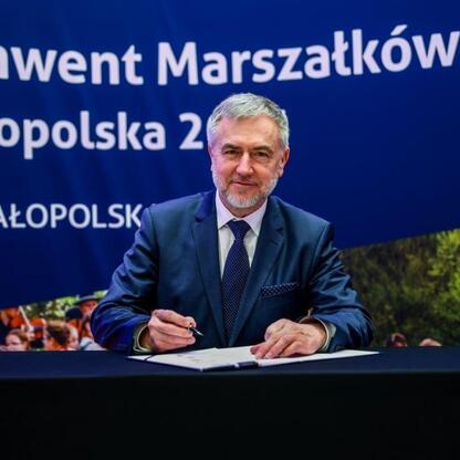 Mężczyzna w garniturze podpisuje dokument na konwencie Marszałków Województw Małopolska 2023. (wygenerowano przez AI)