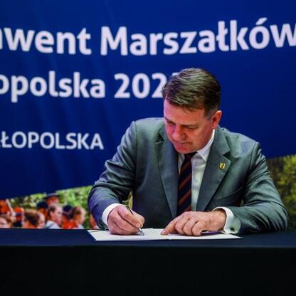 Mężczyzna w garniturze podpisuje dokument na konwencji Marszałków Województw Małopolska 2023. (wygenerowano przez AI)