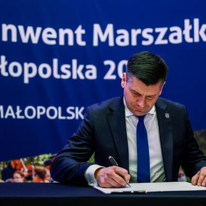 Mężczyzna w garniturze podpisuje dokument, podczas gdy druga osoba stoi za nim. Tło zaznacza Konwent Marszałków Małopolska 2023. (wygenerowano przez AI)