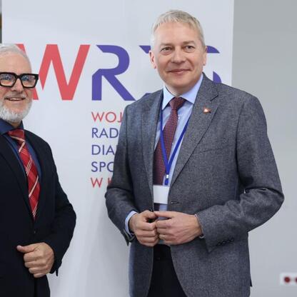 Dwa mężczyźni w strojach formalnych stoją przed tłem z napisami 