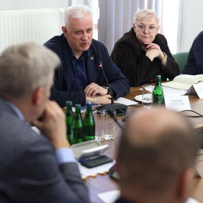 Podpis pod zdjęciem: Grupa ludzi w ścisłym stroju biznesowym siedzi wokół stołu konferencyjnego, prowadząc dyskusję podczas briefingu prasowego. Atmosfera formalna, z mikrofonami, tablicami imiennymi  (wygenerowano przez AI)