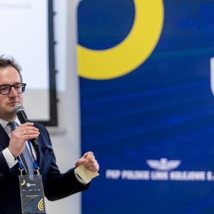 Mężczyzna w garniturze przemawia na podium, gestykulując, podczas warsztatów dotyczących Zintegrowanej Sieci Kolejowej w Województwie Śląskim. (wygenerowano przez AI)
