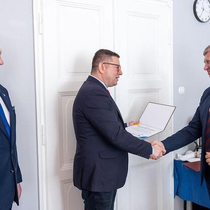Trzech mężczyzn w strojach formalnych w sali konferencyjnej, rozmawiających i podpierających się dokumentem, podczas spotkania Wojewódzkiej Rady Rynku Pracy. (wygenerowano przez AI)