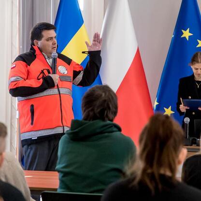 Konkurs wiedzy o pierwszej pomocy województwa Śląskiego. Uczestnik wskazuje na flagi, a publiczność słucha. (wygenerowano przez AI)