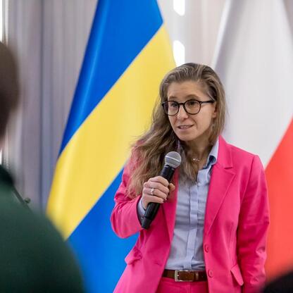 Kobieta w różowej kurtce przemawia do mikrofonu, podczas finału konkursu wiedzy o pierwszej pomocy organizowanego przez Śląskie Youth Parliament. (wygenerowano przez AI)