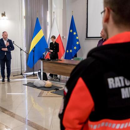 Wojewódzka konkurs wiedzy o pierwszej pomocy, Rafał Adamczyk przemawia, sędzia i uczestnik w mundurze medycznym. (wygenerowano przez AI)
