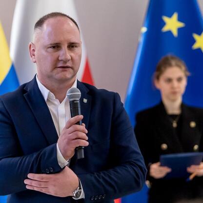 Mężczyzna w ciemnym garniturze przemawia do mikrofonu, tło: flagi Ukrainy i UE, konferencja, Silesia. (wygenerowano przez AI)